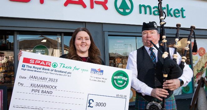 woman with cheque and man with bagpipes in front of Spar store